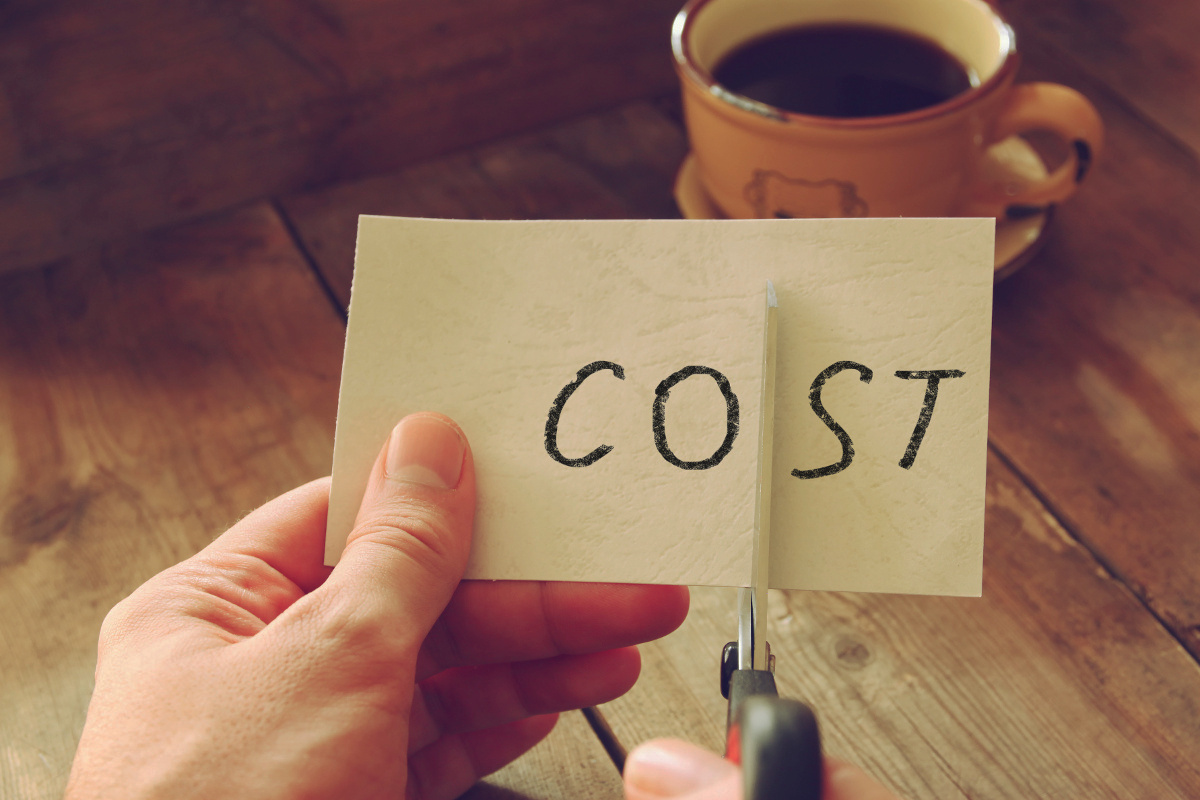 Hands Using Scissors to Cut Paper with the Word 'Cost' on Blurred Background with Cup of Coffee